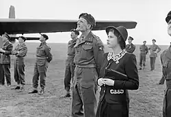 Photo en noir et blanc. Des hommes en uniforme et une femme en tailleur noir (la reine) observent le ciel, sur un terrain herbeux.