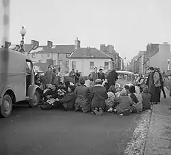 Photo en noir et blanc d'une quinzaine de personnes assises sur une route, empêchant voitures et camions de passer, avec d'autres personnes debout aux alentours.