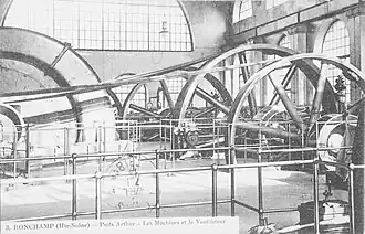 Photo noir et blanc montrant une  vaste salle lumineuse, les grandes roues des machine à vapeur actionnent de grands ventilateurs en forme d'escargot.