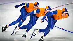 Trois patineurs en combinaison orange, l'un à la suite de l'autre, en fin de virage.