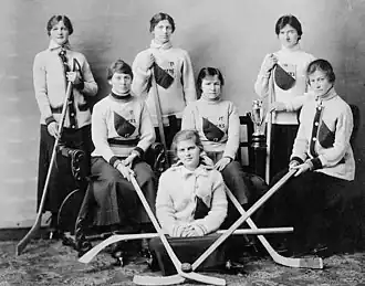 Photo en noir et blanc d'un groupe de femmes en jupe longue et blouse blanche, posant avec des crosses de hockey.