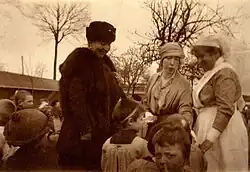 à gauche Henriette vêtue d'un manteau et d'une toque en fourrure souriante regarde un enfant qui reçoit un présent de la reine Élisabeth coiffée d'un bandeau aux côtés d'une infirmière souriante de profil