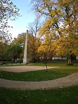 square avec pelouse, allée sablée et arbres au feuillage doré d'automne. Au centre un obélisque (1738)