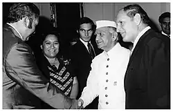 Photo en noir et blanc. Deux hommes se serrent la main devant une femme qui regarde en direction de l'objectif. Un autre homme se trouve à droite de l'image.