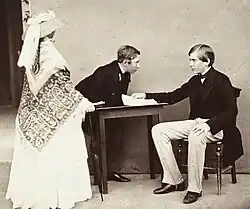 la reine Victoria debout et de dos, vêtue d'une toilette claire, recouverte par un châle brodé et coiffée par un chapeau de paille entouré d'une étoffe nouée regarde ses deux cousins, Louis semi-incliné sur une table regardant son frère Pierre assis et tenant quelques feuilles de papier, les deux princes portent une redingote de couleur foncée