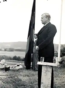 Un homme debout en extérieur derrière un pupitre en bois. Derrière lui, un homme se tient près d'un immense drapeau norvégien.