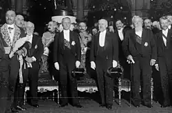 Photo en noir et blanc sur laquelle figurent notamment quatre individus en queue-de-pie se tenant debout