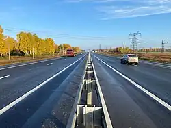 Photographie prise depuis le milieu de la route, avec deux voies de chaque côté, séparées par un séparateur. Du côté gauche, la route est bordée par des mélèzes de Sibérie orangés, car ils vont perdre leurs aiguilles, tandis qu'à droite des pylônes électriques s'élèvent sur un terrain déboisé.