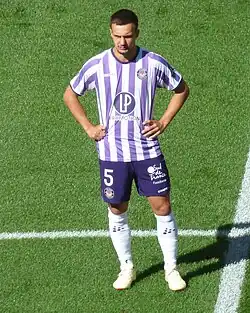 Photo en pied de Denis Genreau, avec les couleurs blanches et violettes de Toulouse sur un terrain de football.