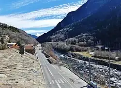 Photographie d'une route franchissant une agglomération avec des maisons aux toits de pierres et une rivière agitée à droite ; on aperçoit un rond-point tout au fond ainsi que des passages piétons et parkings.