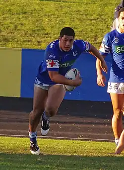 Photographie d'un homme portant un maillot de rugby bleu, tenant un ballon de rugby.