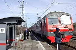 Un long train de passager, peint en rouge et gris, traverse le milieu de l'image. Les wagons sont rouges, et les portes sont ouvertes. Le train est arrêté en gare, la photo est prise depuis le quai juste devant la locomtive électrique, et à gauche, une petite aubette, typique des zones rurales, se distingue. Des fils électriques, soutenus par des pylônes, parallèles au train, courent vers l'avant-plan, tandis qu'à l'arrière-plan, une forêt de conifères sombres semble border la voie. Le ciel est dégagé mais la lumière est rasante.
