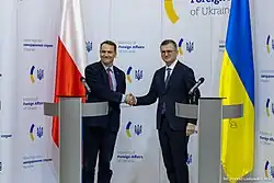 Photo de deux hommes politiques, en costard-cravate, se serrant la main devant un drapeau polonais et un drapeau ukrainien. Kouleba se tient à droite de la photo devant le drapeau ukrainien.