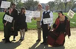 Photo de groupe de six personnes en costume, portant des écriteaux avec des slogans en rapport avec la série