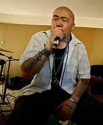 Homme au crâne rasé, aux bras tatoués, chantant dans un micro devant des musiciens avec des instruments traditionnels