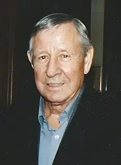 Portrait d'un homme âgé aux cheveux gris portant une veste noire sur une chemise bleue.