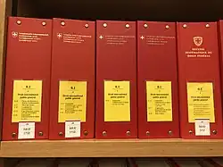 Photographie de cinq classeurs rouges avec étiquette jaune contenant le Recueil systématique sur une étagère en bois