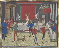 Enluminure médiévale représentant un banquet. Les serviteurs portent de longues chaussures. Deux des dames attablées sont coiffées d'un hennin.