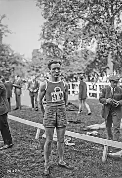 Photographie en noir et blanc d'un homme en short se tenant debout, les mains derrière le dos. Il porte le dossard no 99.