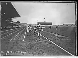 Photographie en noir et blanc d'un homme en tenue de sport, franchissant une ligne d'arrivée. Il s'agit de René Wiriath. Il porte le dossard no 90. En arrière-plan, un groupe de 3 coureurs se présente sur la ligne d'arrivée.