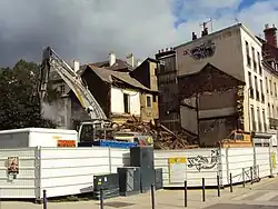Un engin de chantier, derrière des palissades métalliques, procède à la destruction d'un bâtiment.