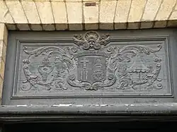 Une photo de l'imposte de la porte principale de l'église. Le blason de la basilique, encadré de ses attributs (pavillon et tintinnabule), est sculpté en bas-relief sur un panneau de bois sombre.