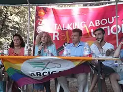 Photographie d'un groupe de personnes attablées dont une femme parlant dans un micro, avec un drapeau LGBT.