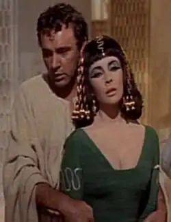 Photographie en couleurs d'une femme en robe verte arborant une riche coiffure, et un maquillage profond sur les paupières, enlacée par derrière par un homme portant une toge claire.