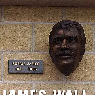 Photo d'un mur de briques sur lequel se détache la tête en bronze d'un homme moustachu aux cheveux courts. Une plaque indique : Robbie James 1957-1998