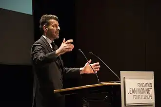 Photographie d'un homme caucasien avec barbe et cheveux gris, parlant à un pupitre.