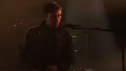 Un homme parle dans un micro lors d'un concert; On peut voir des instruments électroniques en fond.