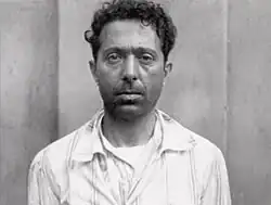 Portrait noir et blanc de face d'un homme brun aux cheveux courts et bouclés portant une chemise blanche, à l'air sérieux.