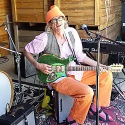 Photo d'un homme blanc âgé aux longs cheveux gris. Il porte un gilet à carreaux noir et blanc, une chemise à rayures roses et un pantalon orange. Assis sur un amplificateur, il joue d'une guitare électrique verte couverte de paillettes.