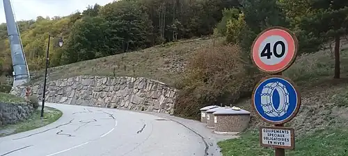 Photographie d'un panneau routier qui indique que la limitation kilométrique est de 40&nbsp;km/h ; un équipement de pneus est nécessaire en hiver ; on voit les conteneurs à ordures pour le tri sélectif derrière, le tout depuis une route.