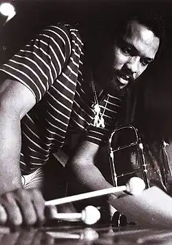 Photo en noir et blanc de Roy Ayers. Vu en contre-plongée, il est penché sur un vibraphone et tient des baguettes dans sa main droite.