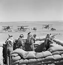 photographie en noir et blanc montrant un groupe de soldat dans une fortification en sacs de sable avec des avions biplan stationnés à l’arrière-plan