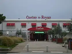 Photographie du Casino de Royan montrant un bâtiment blanc horizontal sur deux niveaux. Le dais marquant l’entrée, les stores de l’étage et l’enseigne sur le toit plat sont de couleur rouge.