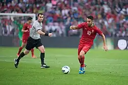 Photo d'un footballeur en tenue rouge sur la droite avec le ballon au sol au centre et l'arbitre observant la scène à gauche.