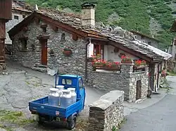 La photo couleur montre un petit triporteur motorisé bleu chargé de 4 bidons de lait en aluminium. La scène est située dans un village aux maisons de pierre et aux toits en dalle de pierre et ardoises.