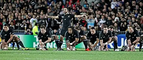De face, plusieurs hommes, en tenue de rugby noire, accroupis. Un autre joueur les bras étendus sur le côté, est au centre, au milieu des des autres.