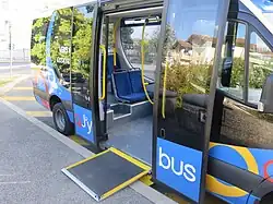 Photographie en couleurs d’un minibus bleu et noir en stationnement avec la rampe d’accès déployée et les portes ouvertes.