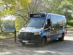 Photographie en couleurs d’un minibus bleu, blanc et noir à proximité d'un espace vert et du plan d’eau de Rumilly, situé en arrière-plan.