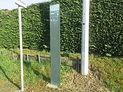 Photographie en couleurs d’un arrêt de bus non couvert : deux panneaux sont dressés sur un accotement en herbe, le tout devant une haie dense en arrière-plan.