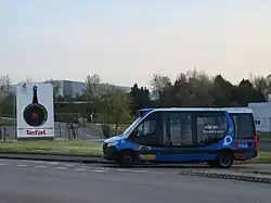 Photographie en couleurs d’un minibus bleu, blanc et noir devant une usine Tefal