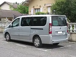 Photographie en couleurs d’une voiture grande capacité grise, garée devant une maison.