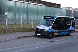 Photographie en couleurs d’un minibus bleu, blanc et noir, stationné devant des bâtiments à l'aspect moderne