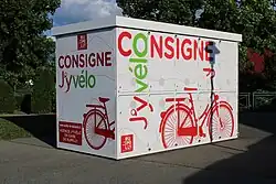 Photographie en couleurs d’une consigne à vélo, un cube blanc installé sur le trottoir et sur lequel figurent plusieurs dessins de vélos.