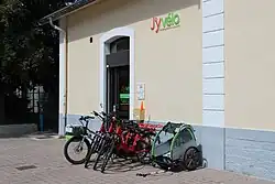 Photographie en couleurs d'un bâtiment de location de vélos J'yvélo devant lequel sont alignés plusieurs vélos.