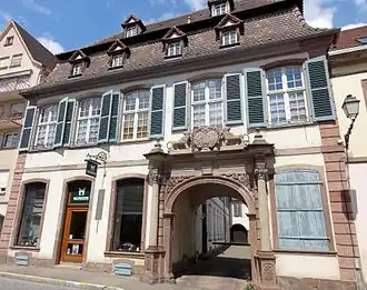 Photographie d'un bâtiment blanc de deux étages avec une arche sculptée en gré rose qui donne sur une cour intérieure.
