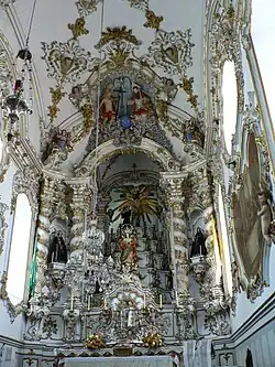 Photographie en couleurs. Dans un intérieur d'église très éclairé, un grand retable blanc avec des décorations dorées s'inscrit dans un cadre de la même configuration.